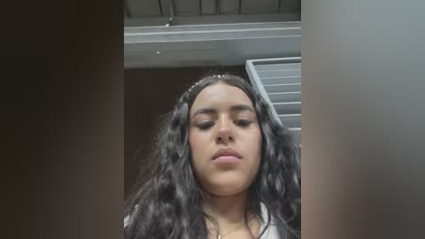 Media: A video of a young, light-skinned woman with long, wavy black hair, wearing a white T-shirt, captured from a low angle, with a ceiling fan and part of a wall visible in the background.