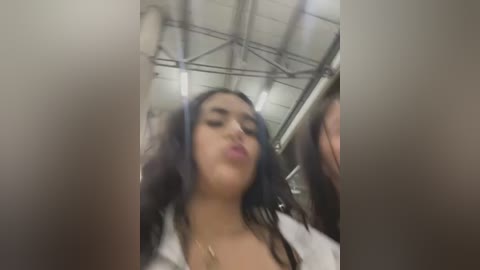 Media: A blurry, low-resolution video of a young woman with long, dark hair, wearing a white top, in a warehouse setting with exposed beams and industrial lighting.