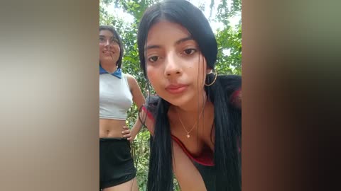 Media: Video of two women in a lush green forest. One woman, with long black hair, wears a black top and black shorts, while the other, with dark hair, wears a white crop top and black shorts.