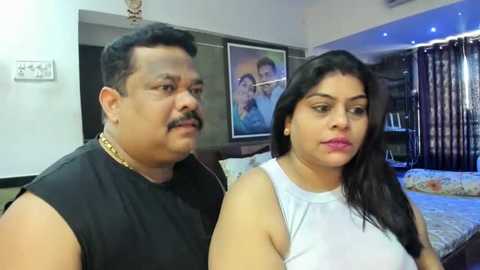 Media: Video of an overweight, bearded man in a black shirt and a plus-sized woman with long black hair in a white top, both indoors, near a bed with floral bedding, framed pictures on the wall.