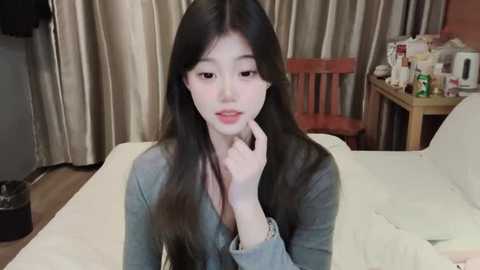 Media: A video of a young East Asian woman with long, straight black hair, wearing a gray cardigan, sitting on a bed with white sheets. Background features beige curtains, a wooden chair, and a cluttered table with various items.