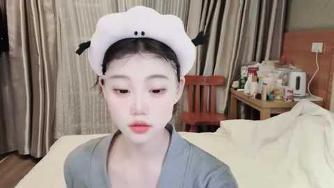 Media: Video of a young Asian woman with pale skin, wearing a white hat resembling a dog's head and a blue dress, sitting on a bed in a bedroom with beige curtains, wooden furniture, and a cluttered table.