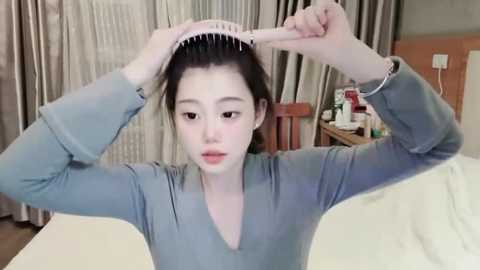 Media: A video of an East Asian woman with fair skin and dark hair in a grey sweater, combing her hair in a cozy room with wooden furniture and beige curtains.