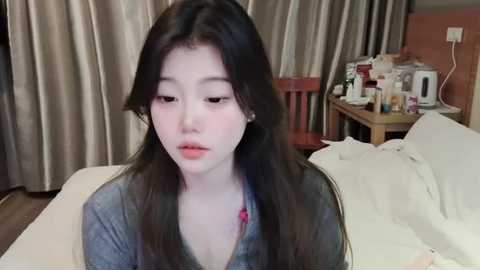 Media: A video of a young East Asian woman with long black hair, fair skin, and a soft expression, sitting on a bed in a dimly lit room with beige curtains and a cluttered nightstand.