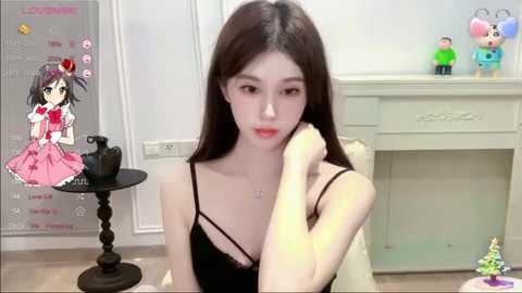 Media: A video of an Asian woman with long black hair, fair skin, and a slender physique, dressed in a black camisole, sitting indoors with a pink dress overlay, surrounded by toys.