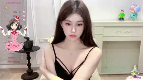 Media: A video of an Asian woman with long dark hair, fair skin, and light makeup, wearing a black lace camisole, sitting on a cream couch, in a minimalist room with white walls and a black side table.