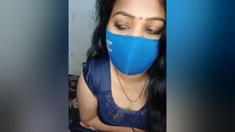 Media: A video of a South Asian woman with medium skin tone, wearing a blue mask, black hair, and a blue lace top, with a floral background.