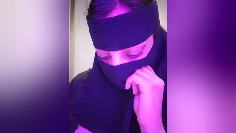 Media: A video of a woman with light skin, wearing a black headband, partially covering her face with a black cloth, set against a purple background.