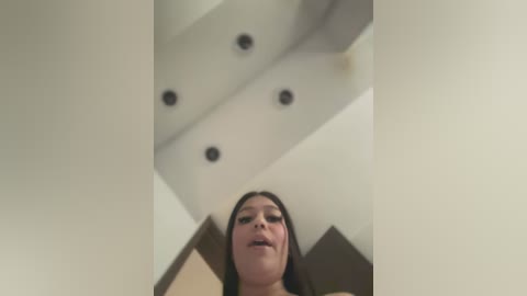 Media: A video of a woman with long dark hair, captured from a low angle, showing her partially obscured face. The background features a modern, minimalist room with geometric ceiling design and recessed lighting.