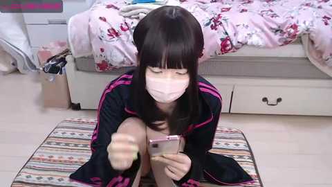 Media: Video of an Asian woman with long black hair, wearing a black jacket with pink stripes, a face mask, and pink headphones, sitting on a striped rug in a room with a white bed and pink floral bedding.