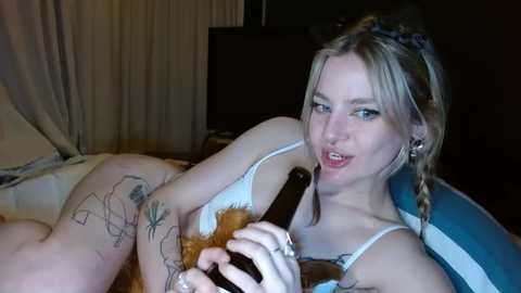 Media: Video of a smiling, blonde woman with braids, wearing a white bra, lying on a bed with a tattoo on her thigh, holding a bottle, dimly lit room.