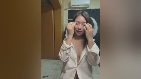 Media: Video of an Asian woman with long black hair, wearing a white button-down shirt, playfully covering her eyes. Background shows a beige carpet, wooden doors, and an air conditioning unit.