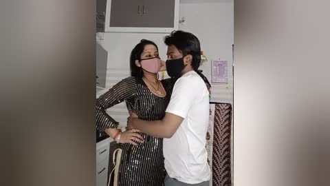 Media: Video of an intimate moment between a South Asian woman with medium-length black hair, wearing a black and white striped dress, and a man with a black mask, white t-shirt, and black hair, in a domestic setting.