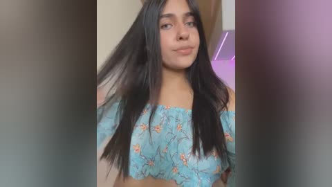 Media: Video of a young woman with long, straight black hair, fair skin, and brown eyes, wearing a light blue off-shoulder top adorned with a floral pattern.