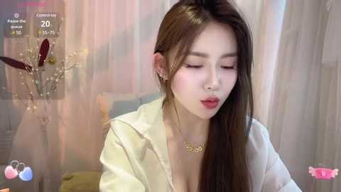 Media: Video of a young Asian woman with long brown hair, wearing a white blouse, and a gold necklace, sitting in a softly lit room with pastel-colored curtains and floral decor.