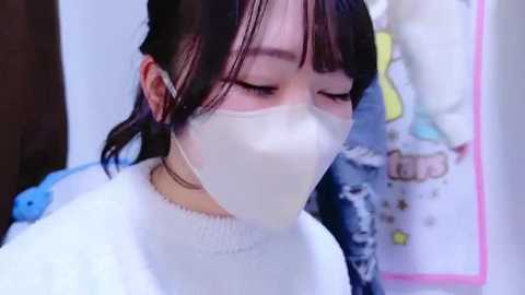 Media: Video of a young East Asian woman with dark hair, wearing a white mask and a white sweater, standing in a room with a wall calendar and stuffed animals in the background.