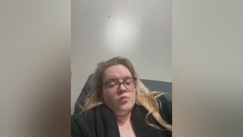 Media: Video of a fair-skinned woman with blonde hair, wearing glasses, lying on a black chair, resting her head on her hand, against a plain, light-colored wall.