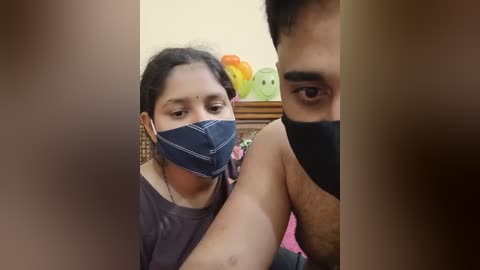 Media: Video of a South Asian woman with dark skin, wearing a blue face mask and a purple shirt, and a man with light skin, wearing a black face mask, in a cozy, colorful room with toys.