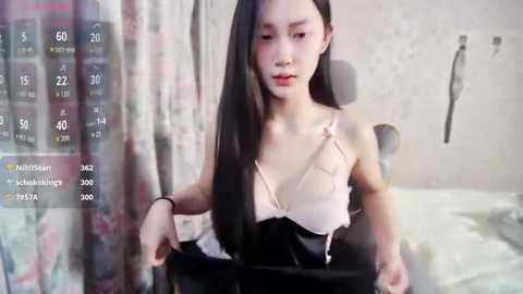 Media: Video of an East Asian woman with long black hair, wearing a pink and black lingerie set, sitting on a bed with a messy background.