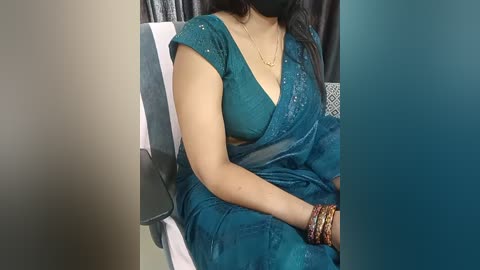 Media: Video of a South Asian woman in a teal saree with a plunging neckline, seated on a black chair, wearing a black mask, with a blurred background.