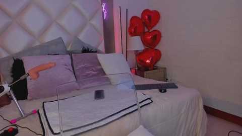 Media: Video of a modern bedroom with a bed, white duvet, grey pillows, heart-shaped helium balloons, and a white nightstand with a lamp. The room features a white quilted headboard and a minimalist, chic design.