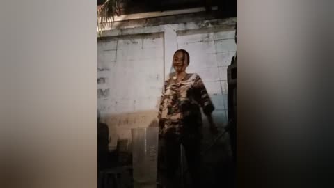 Media: A video captures a middle-aged man with short hair, wearing a camouflage-patterned shirt and pants, standing in a dimly lit, worn-out room with peeling white walls and a cluttered background.