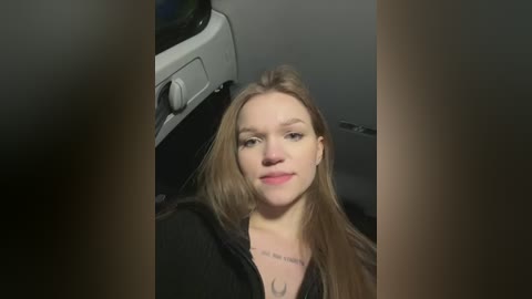 Media: Video of a young Caucasian woman with long, straight, light brown hair, wearing a black top, seated in a car. She has a faint smile and a tattoo on her neck.