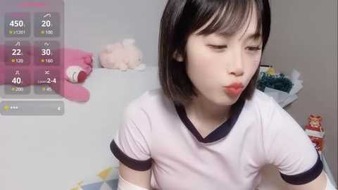 Media: Video of an East Asian woman with fair skin, straight black hair, and a small build, wearing a white T-shirt with black trim, blowing a kiss. Background shows a simple room with a pink teddy bear, a white shelf, and a carton.