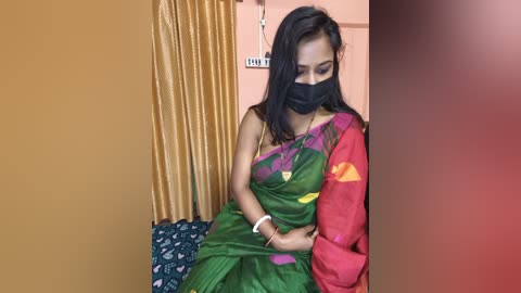 Media: A video of a young South Asian woman with medium skin tone and long black hair, wearing a green sari with pink accents, a black face mask, and a white bracelet, sitting in a room with a mustard-yellow curtain and a red wall.