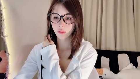 Media: Video of a young East Asian woman with straight, shoulder-length brown hair, wearing black-rimmed glasses and a white hoodie, sitting in a modern bedroom with beige curtains and a bed with white sheets.