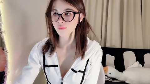 Media: Video of a young, slender, pale-skinned woman with straight brown hair, wearing black-rimmed glasses and a white hoodie, standing in a bedroom with white curtains, a bed, and stuffed animals.