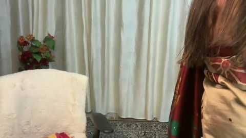 Media: Video of a woman with long brown hair, wearing a red scarf and beige jacket, standing near a white armchair with a grey pillow. Background features white curtains and a floral arrangement with red and orange flowers.