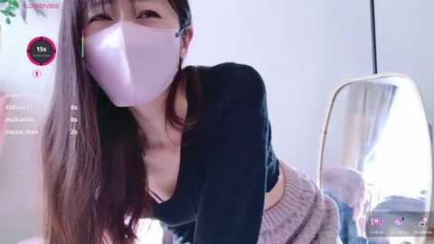 Media: Video of an East Asian woman with long brown hair, wearing a pink face mask, black long-sleeve top, and gray knit skirt, bending over a mirror, in a softly lit room.