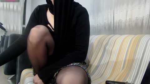 Media: Video of a person, likely a woman, sitting on a striped couch, wearing a black face mask, black top, and short patterned skirt. Background includes a tiled wall and blurred furniture.