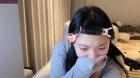 Media: Video of an Asian woman with long black hair styled into two buns, wearing a grey cardigan, covering her mouth with a white cloth, sitting on a white chair in a messy bedroom.