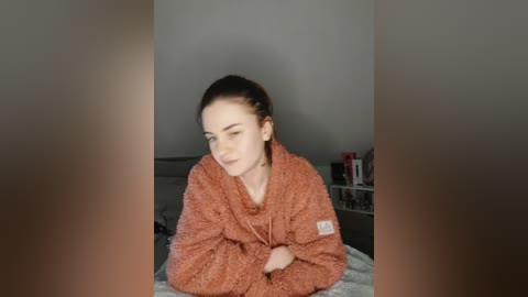 Media: A video of a fair-skinned, young woman with long brown hair in a bun, wearing a light brown, fluffy robe, resting her head on her hands in a dimly lit bedroom with a bed and a cluttered nightstand in the background.