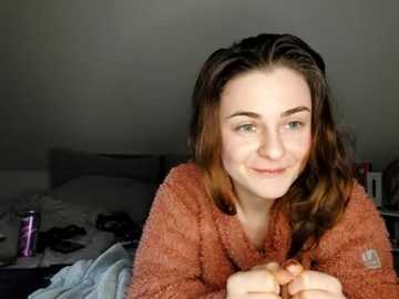 Media: Video of a smiling young woman with wavy brown hair, wearing a cozy brown fleece robe, lying on a messy bed with gray pillows and scattered clothes.