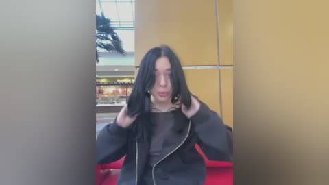 Media: Video of a young woman with long, black hair, wearing a black jacket and black shirt, standing indoors with a blurred background featuring a mall area.