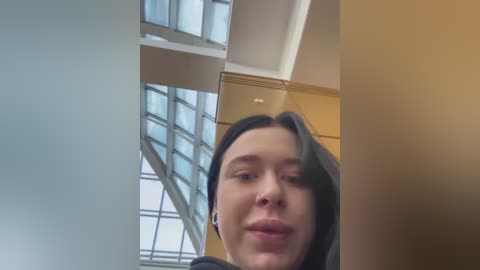 Media: Video of a young woman with straight dark hair, wearing a black top, standing in a modern room with large glass windows and wooden cabinets in the background.