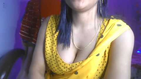 Media: Video of a fair-skinned woman with long blue hair, wearing a low-cut yellow top with polka dots, revealing ample cleavage. She has dangling earrings and a necklace. Background shows a blurred, colorful room with purple lighting.