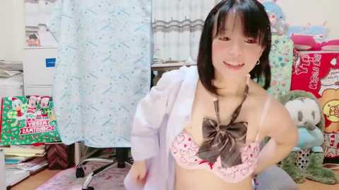 Media: Video of an Asian woman with straight black hair, wearing a pink lace bra with a bow, and a white lab coat, smiling in a cluttered bedroom adorned with anime posters and stuffed animals.