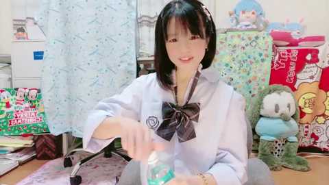 Media: Video of a young Japanese woman with straight black hair, wearing a white shirt with a black bow, smiling while pouring water from a green bottle. Background features colorful, patterned curtains, stuffed toys, and a cluttered desk.