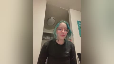 Media: A video of a young woman with shoulder-length, wavy, teal hair, wearing glasses and a black long-sleeve shirt, standing in a hallway with beige walls, a door, and a teal poster.