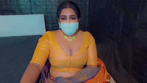 Media: Video of a South Asian woman with dark hair and a light brown complexion wearing a yellow blouse, orange sari, and a blue face mask, sitting on a dark wooden bench in a dimly lit room with dark brick walls.