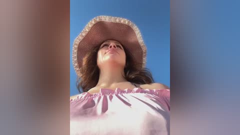 Media: Video of a young woman with fair skin, wearing a large straw hat and pink off-shoulder blouse, gazing upward against a clear blue sky.