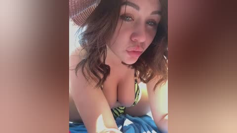 Media: Video of a young woman with wavy brown hair, green eyes, and fair skin, wearing a green and white striped bikini top, lying on a bed with blue sheets, looking sultry into the camera.