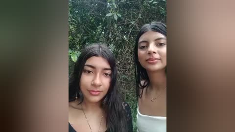 Media: Video of two young women with long black hair, one in a black top, the other in a white off-shoulder top, standing outdoors in front of dense green foliage.