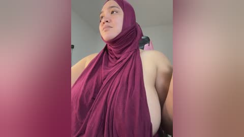 Media: Video of a plus-sized woman with a light brown complexion, wearing a deep magenta hijab and sleeveless top, indoors, blurry background.