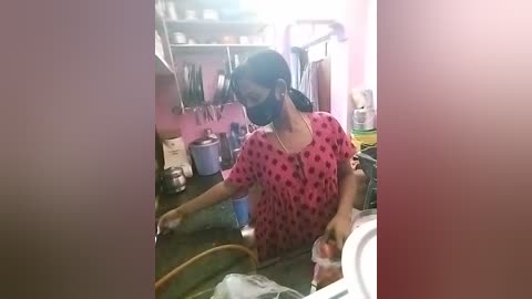 Media: Video of a woman in a pink polka-dot shirt, wearing a mask, kneeling on a dark floor in a cluttered kitchen with shelves and appliances.