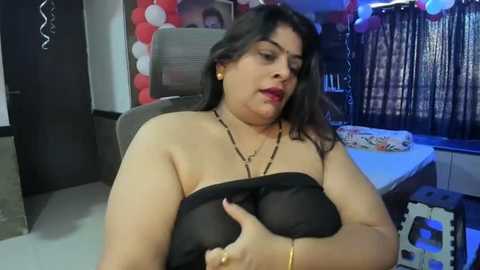 Media: Video of a plus-size woman with medium skin tone, dark hair, and black strapless top, adjusting her breast, in a dimly lit room with balloons and a framed photo.